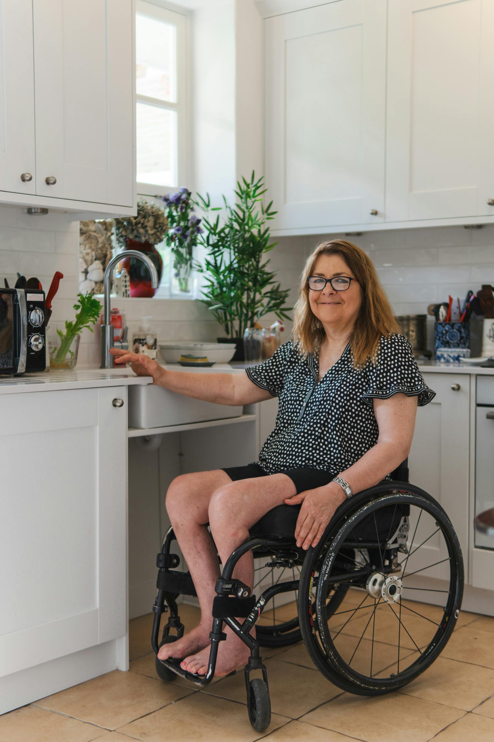 A woman smiling in her wheelchair-accessible kitchen, with the under-sink area open for her to roll up to the counter.